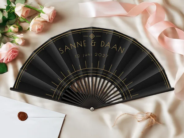 Gepersonaliseerde bruiloftwaaier "Art deco" met de namen Sanne & Daan, fijne bogen en grafische art-decostralen rond het midden, zwarte stof met gouden opdruk, op een bruiloftstafel