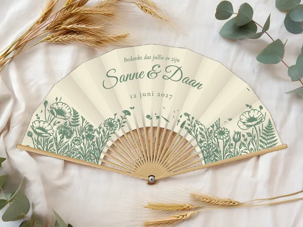 Gepersonaliseerde bruiloftwaaier "Boerentuin" met de namen Sanne & Daan, perk met klaprozen, varens en wilde bloemen eronder, ivoorkleurige stof met saliegroene opdruk, op een bruiloftstafel