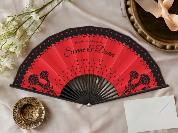 Gepersonaliseerde bruiloftwaaier "Flamenco" met de namen Sanne & Daan, kanten volant met stippen langs de rand en anjers in de hoeken, rode stof met zwarte opdruk, op een bruiloftstafel