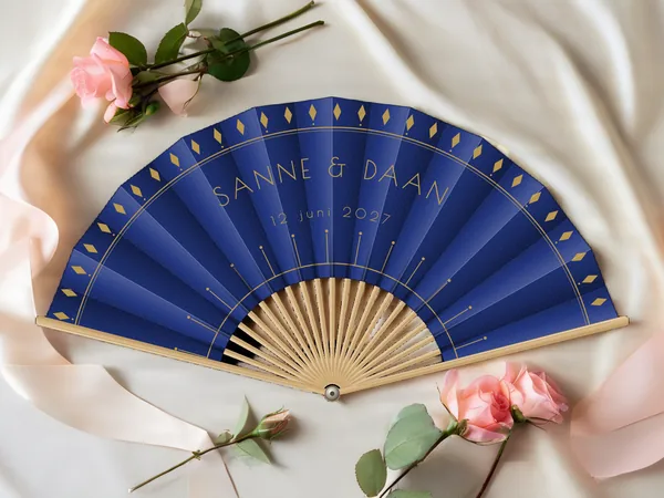 Gepersonaliseerde bruiloftwaaier "Gatsby" met de namen Sanne & Daan, ruiten langs de rand en art-decostralen eronder, marineblauwe stof met gouden opdruk, op een bruiloftstafel