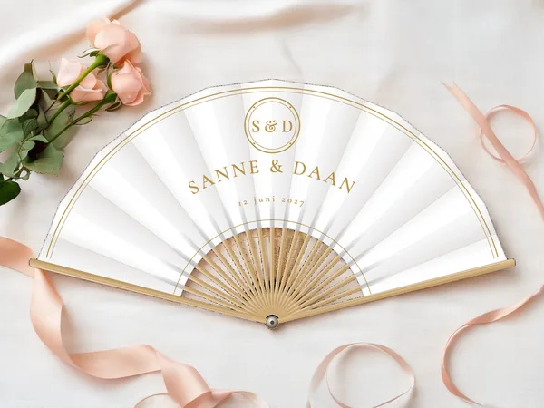 Gepersonaliseerde bruiloftwaaier "Gouden monogram" met de namen Sanne & Daan, medaillon met initialen erboven en een dubbele lijn langs de rand, witte stof met gouden opdruk, op een bruiloftstafel