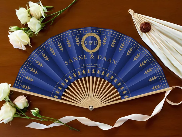 Gepersonaliseerde bruiloftwaaier "Koninklijke laurier" met de namen Sanne & Daan, lauwerkrans erboven en rijen takjes, marineblauwe stof met gouden opdruk, op een bruiloftstafel