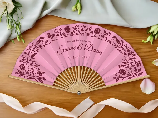 Gepersonaliseerde bruiloftwaaier "Lentebloesem" met de namen Sanne & Daan, slinger van bloemen, vogels en vlinders eromheen, roze papier met bordeauxrode opdruk, op een bruiloftstafel