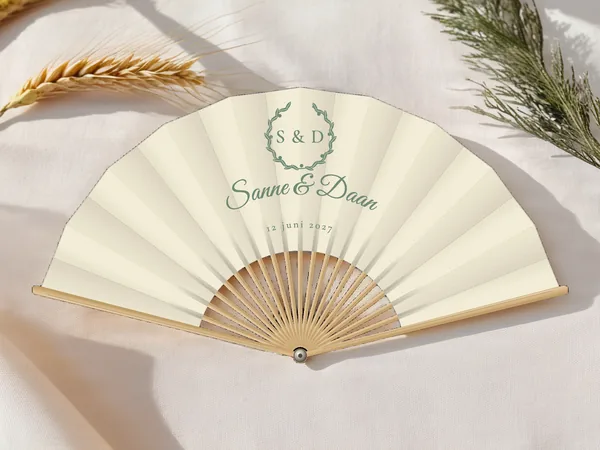 Gepersonaliseerde bruiloftwaaier "Olijfkrans" met de namen Sanne & Daan, bladerkrans rond de initialen erboven, ivoorkleurige stof met saliegroene opdruk, op een bruiloftstafel