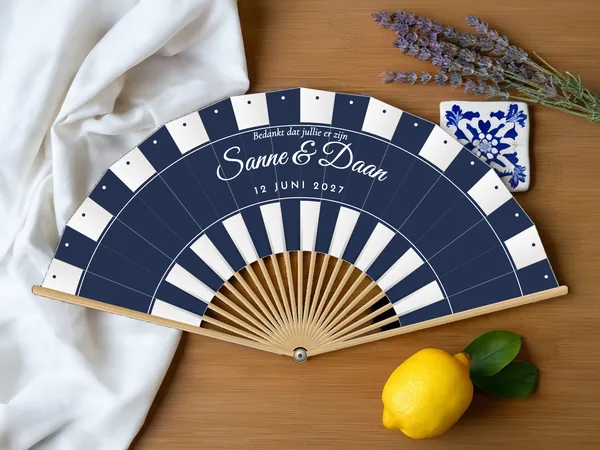 Gepersonaliseerde bruiloftwaaier "Parasol" met de namen Sanne & Daan, parasolstrepen in banen langs de rand en onderaan, witte stof met marineblauwe opdruk, op een bruiloftstafel
